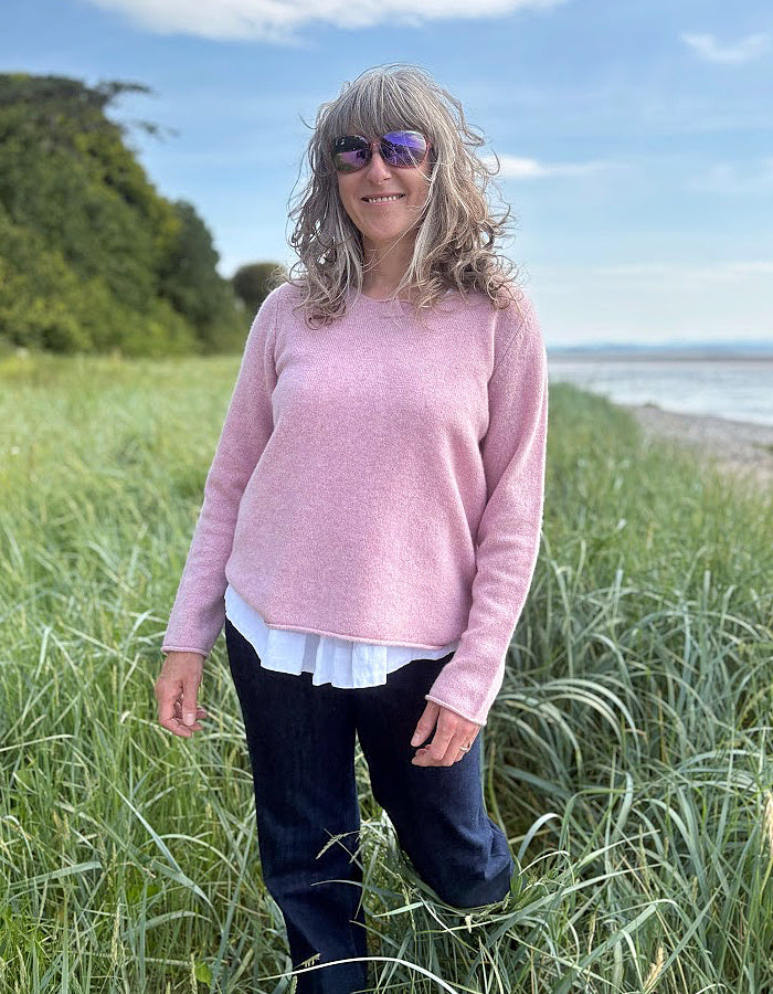 Woman in a pink wool sweater standing in tall grass by a coastal area