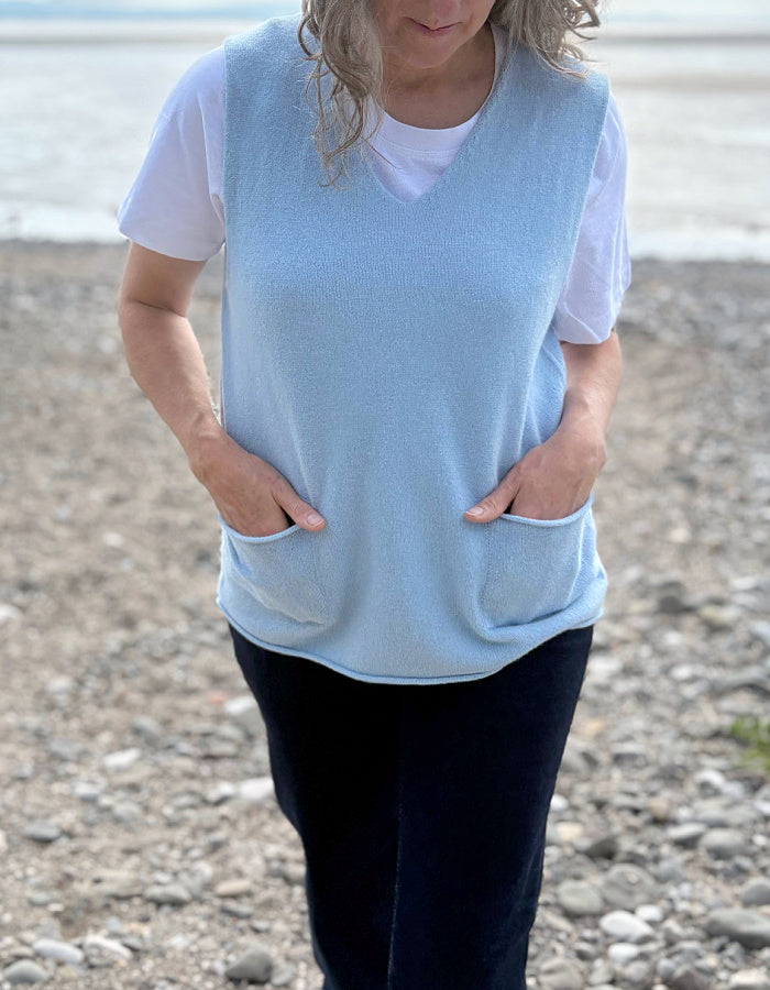 woman wearing a light blue sleeveless top with pockets on a pebbly beach.