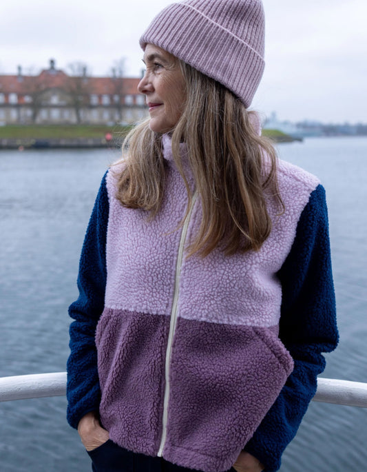 A woman wearing a color-block fleece vest and matching knit beanie stands outdoors by a body of water, looking to the side. The vest has a light lavender upper section, a darker mauve-purple lower section, and navy sleeves, with a light zipper.