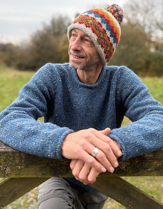 Man wearing a colorful knit hat and blue sweater leaning on a wooden fence outdoors.