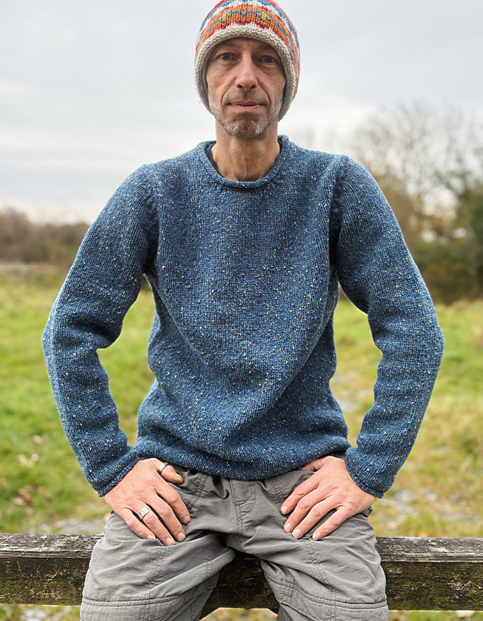 Man wearing a blue knitted sweater and colorful beanie standing outdoors with a natural background.