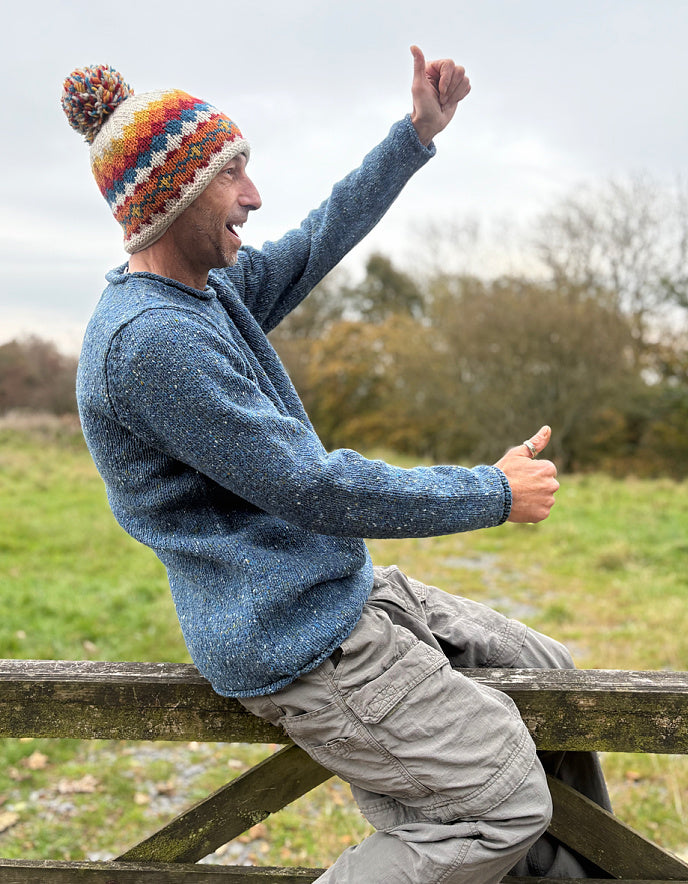Person wearing a blue sweater and colorful knit hat sitting on a wooden fence outdoors.