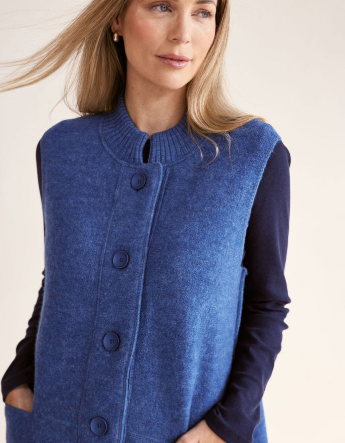 Woman wearing a blue boiled wool long vest over a dark long-sleeve shirt on a light pink background