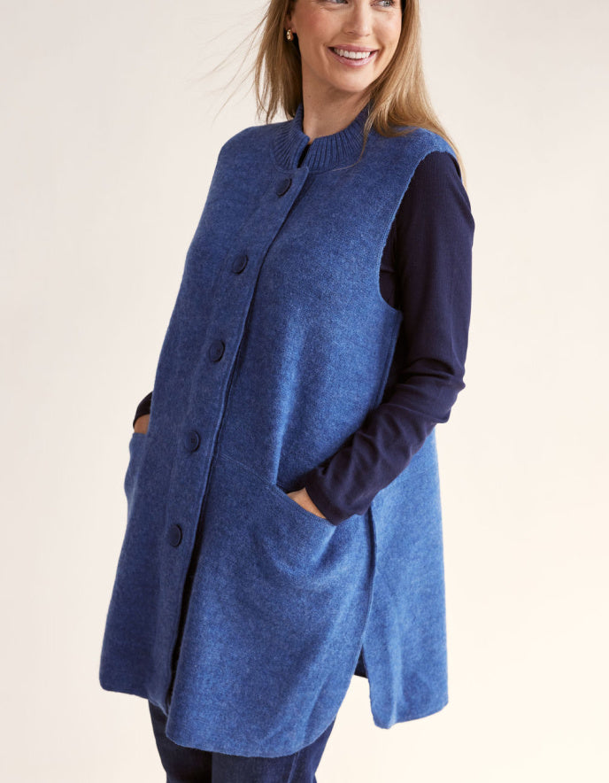 Woman wearing a blue boiled wool long vest over a dark long-sleeve shirt on a light pink background