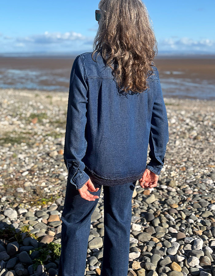 A person standing on a pebble beach, viewed from behind, wearing a dark blue denim long-sleeve shirt and matching denim pants, with water and sky in the background.