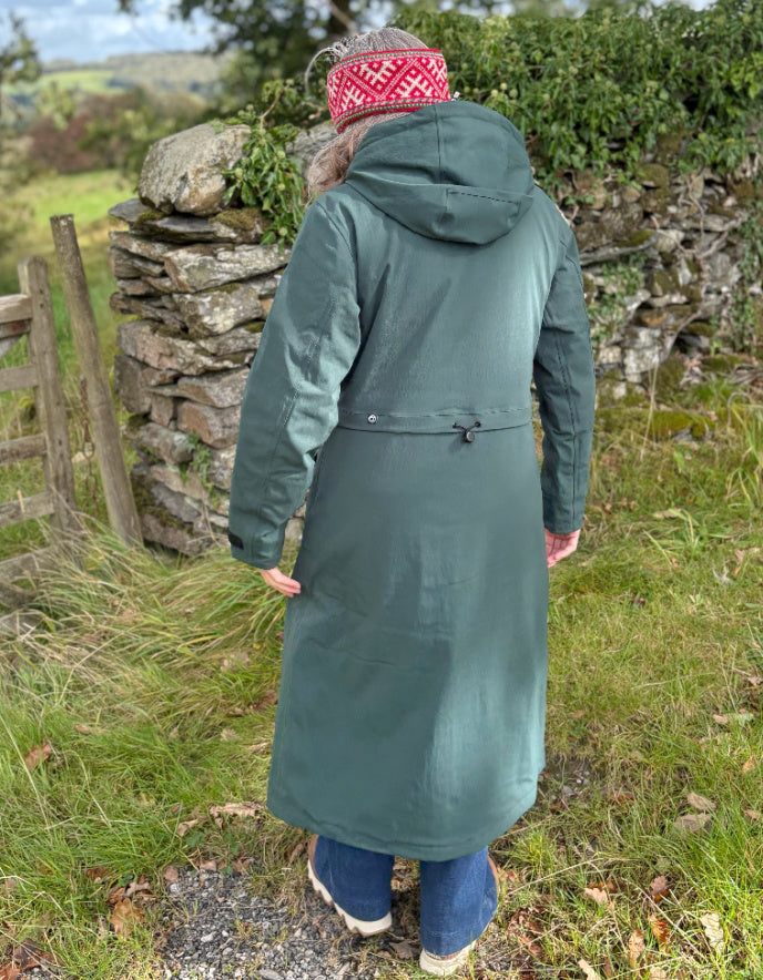 Person wearing a long green coat and patterned headband standing in a natural setting with stone walls and greenery.