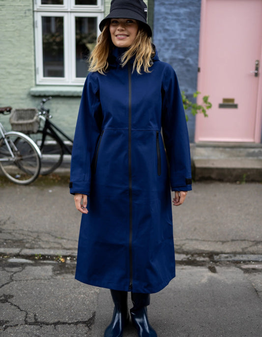 A woman wearing a long navy blue raincoat, a black bucket hat, and dark rain boots stands outdoors on a street in front of a building with a pink door and a bicycle.