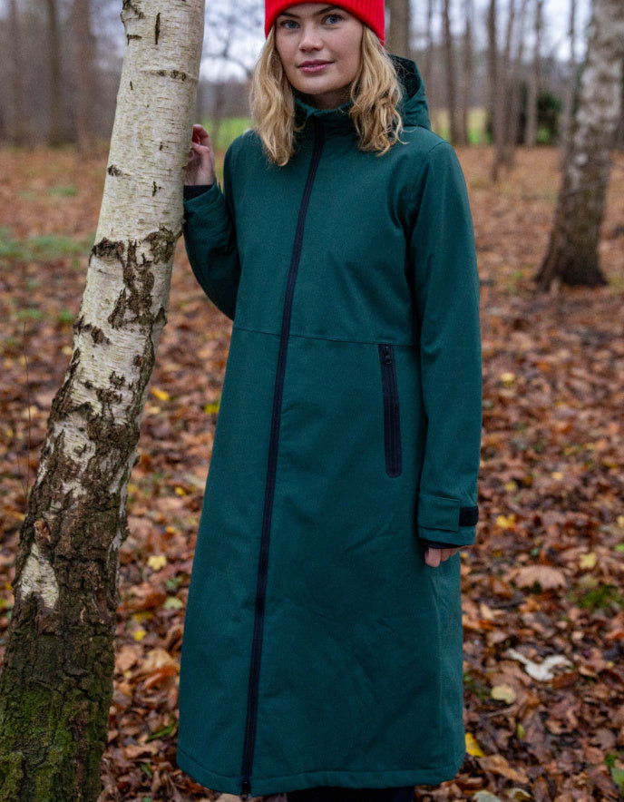 Woman wearing a long teal coat standing in a forest with fallen leaves on the ground.