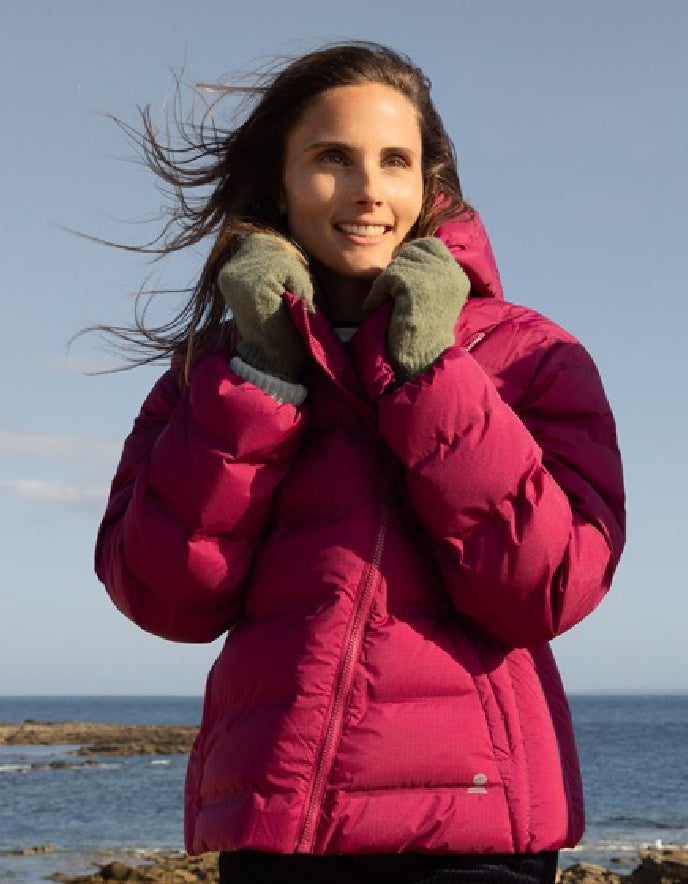 Woman wearing a pink puffer jacket and green gloves standing on a rocky shoreline with ocean and sky in the background.