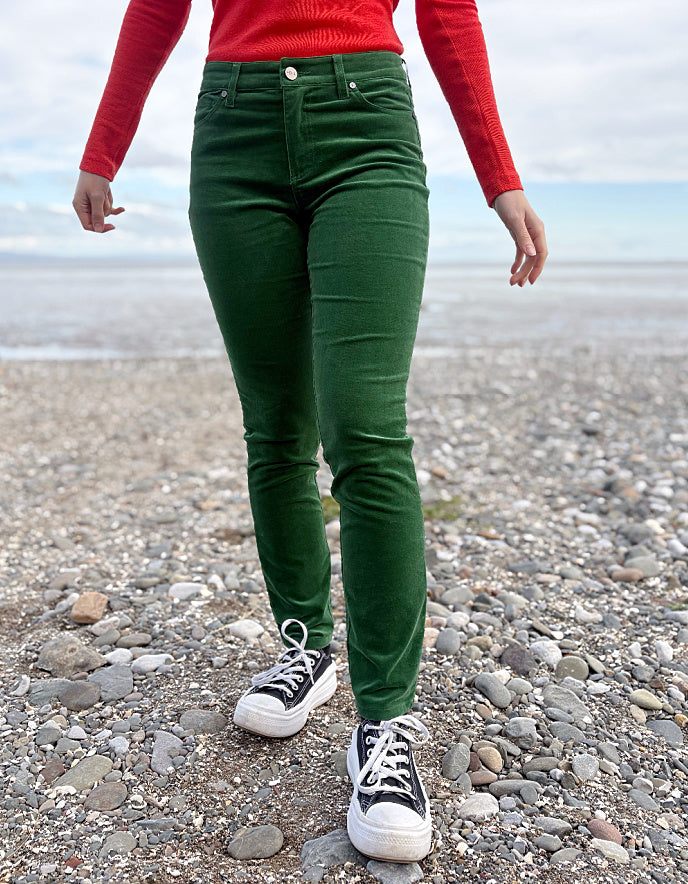 Person wearing green velvet pants and red sweater standing on a pebbly beach.