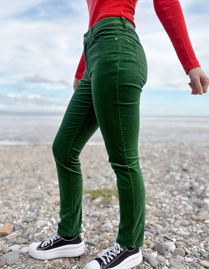 Person wearing green velvet pants and red sweater standing on a pebbly beach.