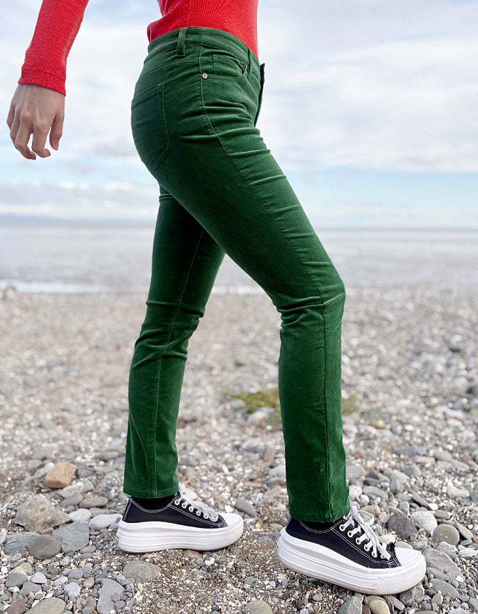 Person wearing green velvet pants and red sweater standing on a pebbly beach.