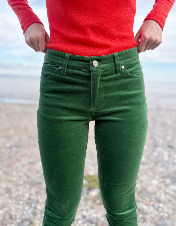 Person wearing green velvet pants and red sweater standing on a pebbly beach.