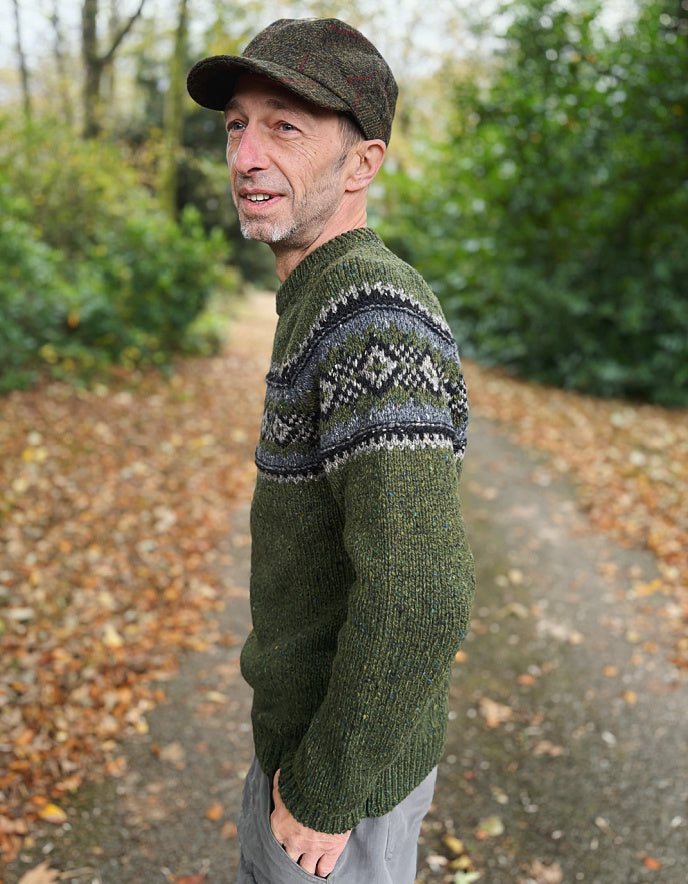 Man wearing a green sweater with a patterned design outdoors on a path with trees and leaves.