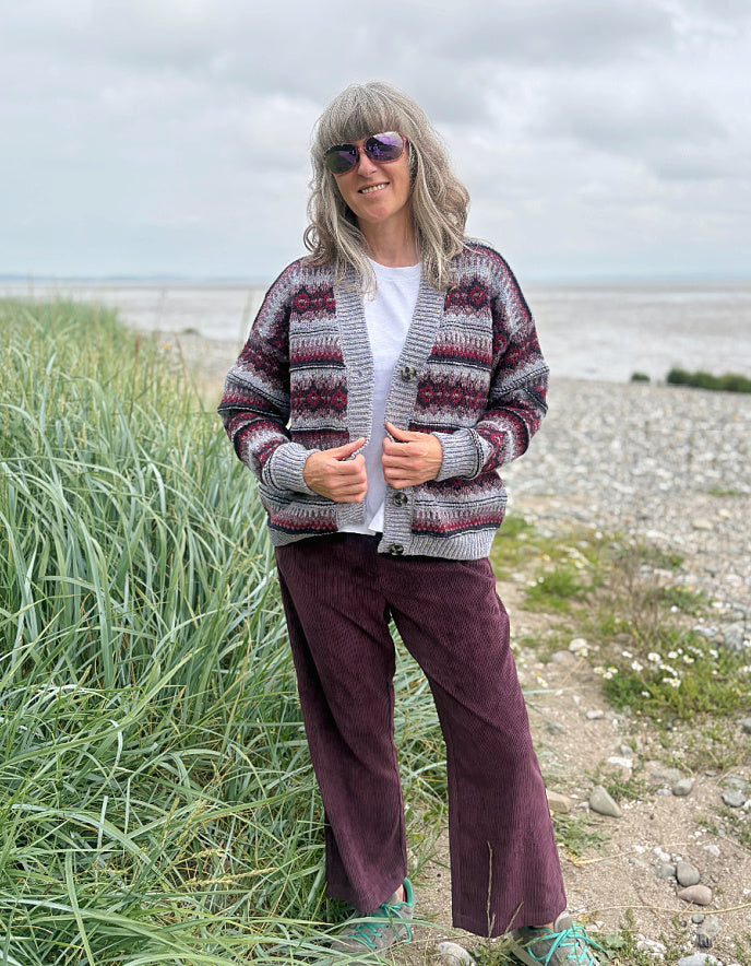 Woman wearing a fair Isle cardigan and plum cord trousers and sunglasses standing by a beach.