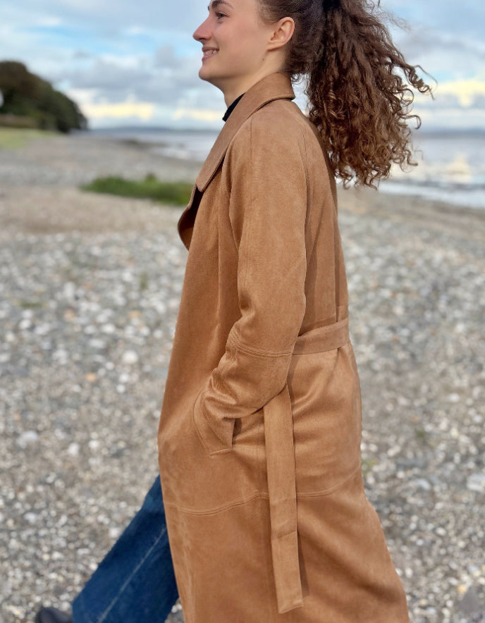 Person wearing a camel faux suede trench coat standing on a beach with a cloudy sky.