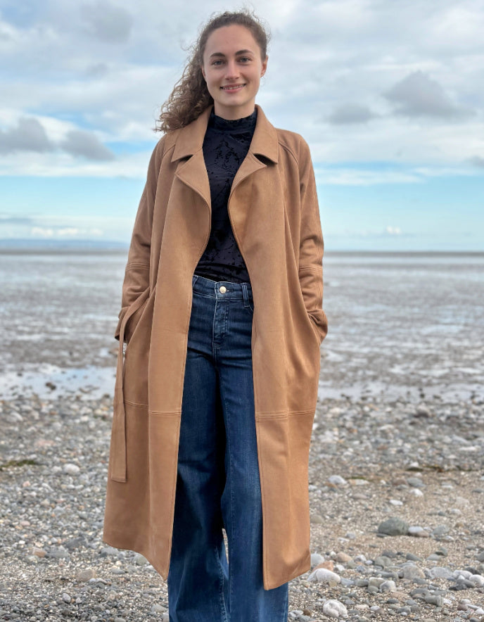 Person wearing a camel faux suede trench coat standing on a beach with a cloudy sky.