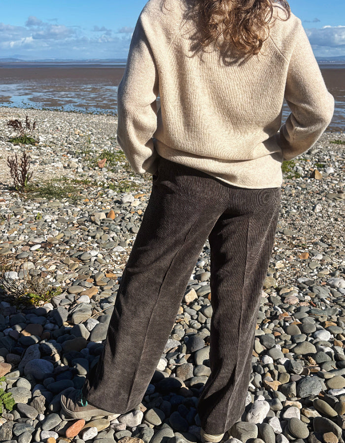 A woman standing on a pebble beach wearing dark brown wide-leg corduroy pants, a cream knit sweater, and dark sneakers, with water and sky in the background.