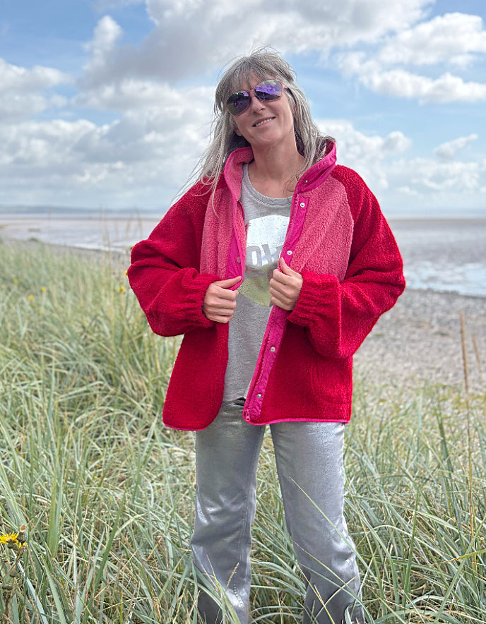 Person wearing a red and pink fleece jacket with silver wide leg jeans standing in grass with a blue sky and clouds in the background