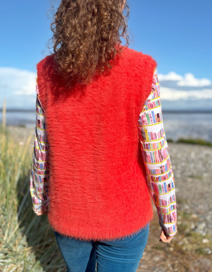 Person wearing an orange fluffy knitted vest over a colourful patterned shirt on a beach.