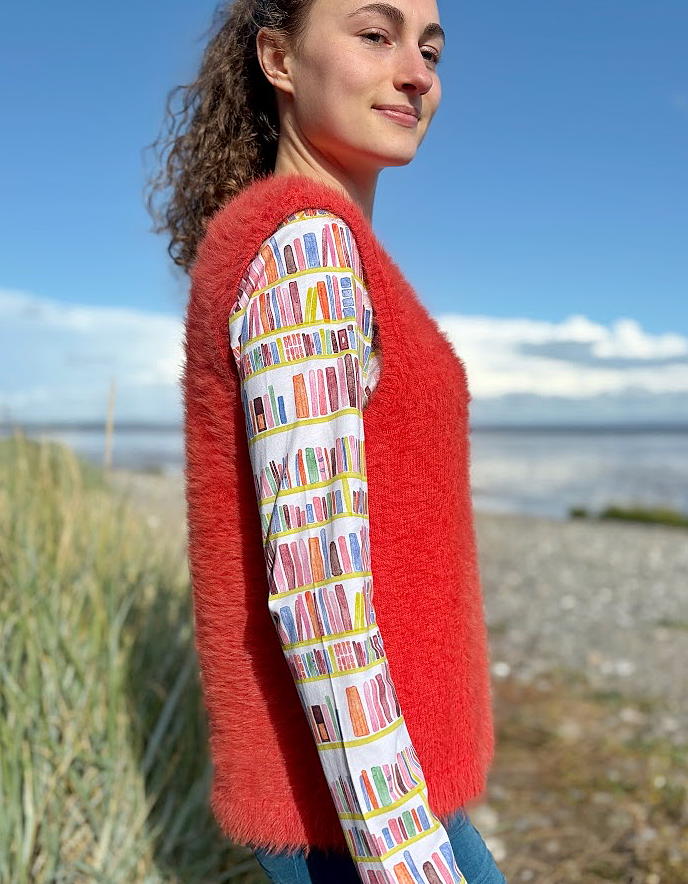Person wearing an orange fluffy knitted vest over a colourful patterned shirt on a beach.