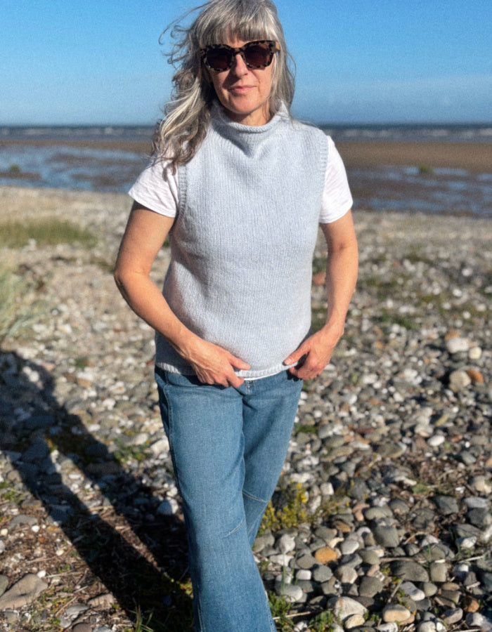A woman standing on a rocky beach wearing a light gray ribbed knit mock-neck sweater vest over a white t-shirt and blue jeans.