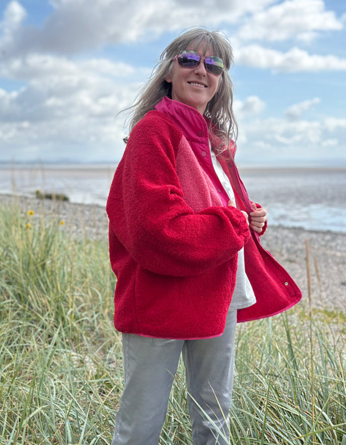 Person wearing a red and pink fleece jacket with silver wide leg jeans standing in grass with a blue sky and clouds in the background