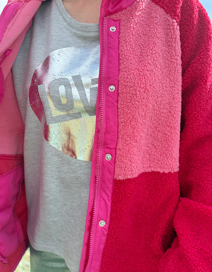 Person wearing a red and pink fleece jacket with silver wide leg jeans standing in grass with a blue sky and clouds in the background