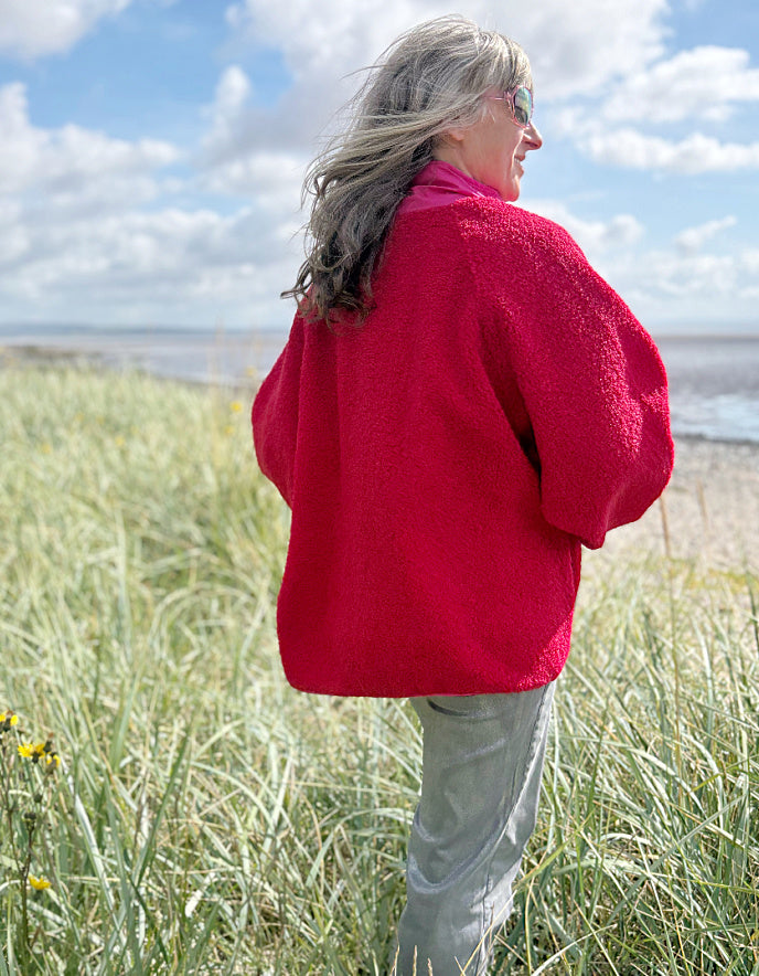 Person wearing a red and pink fleece jacket with silver wide leg jeans standing in grass with a blue sky and clouds in the background