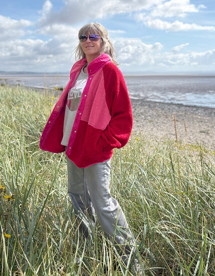 Person wearing a red and pink fleece jacket with silver wide leg jeans standing in grass with a blue sky and clouds in the background