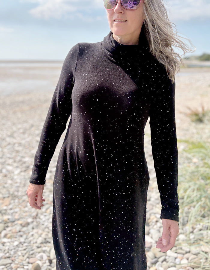 Person wearing a black dress with white speckled galactic print standing on a pebbly beach with a blue sky.