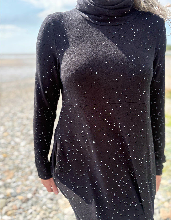 Person wearing a black dress with white speckled galactic print standing on a pebbly beach with a blue sky.