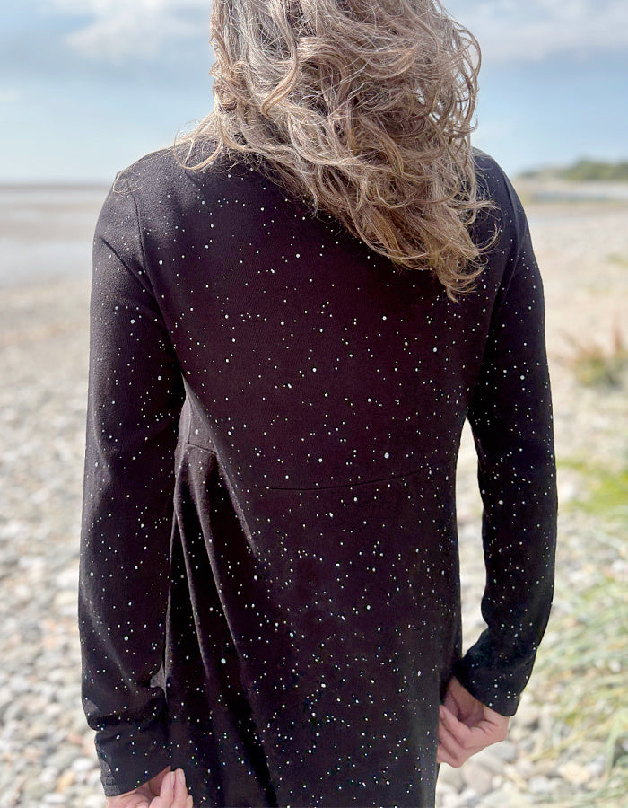 Person wearing a black dress with white speckled galactic print standing on a pebbly beach with a blue sky.