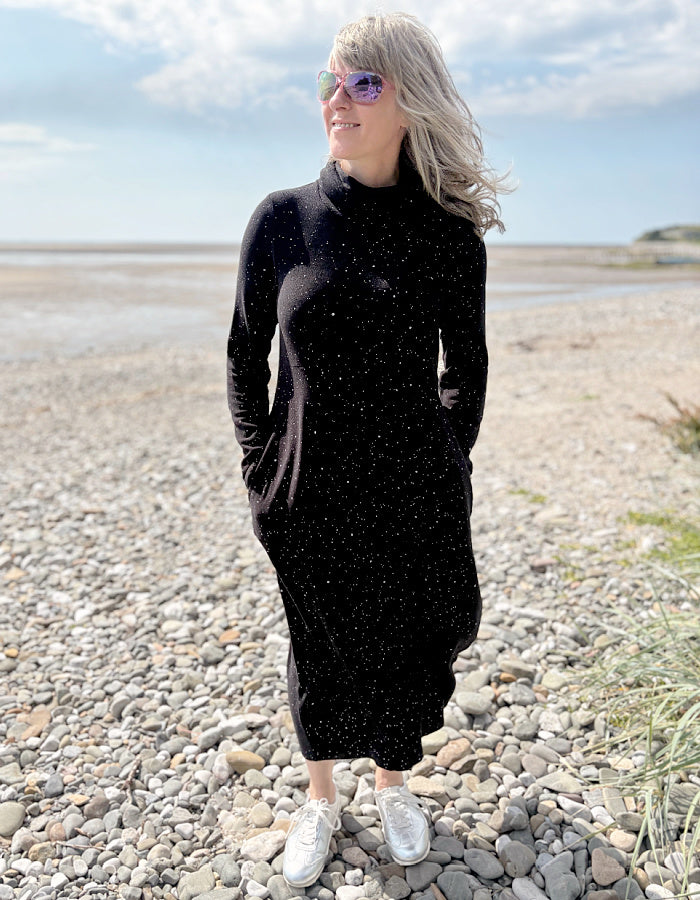 Person wearing a black dress with white speckled galactic print standing on a pebbly beach with a blue sky.