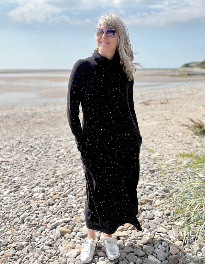Person wearing a black dress with white speckled galactic print standing on a pebbly beach with a blue sky.