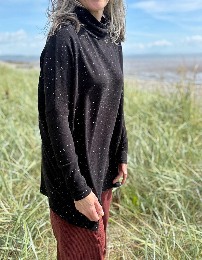 Person standing in a grassy field with a scenic background wearing a black cosy turtle neck tunic and copper brown cord trousers