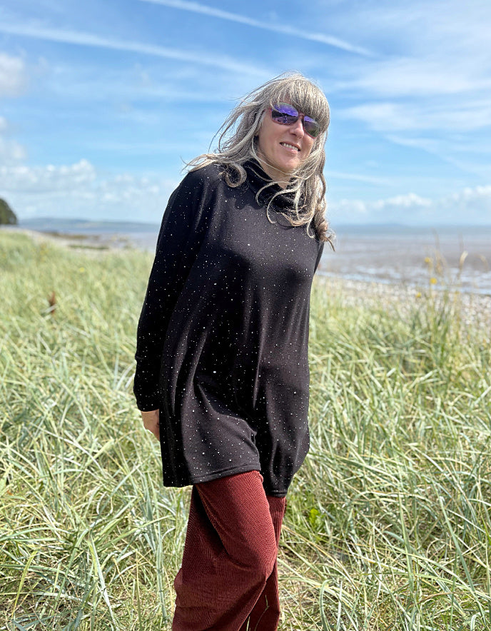 Person standing in a grassy field with a scenic background wearing a black cosy turtle neck tunic and copper brown cord trousers