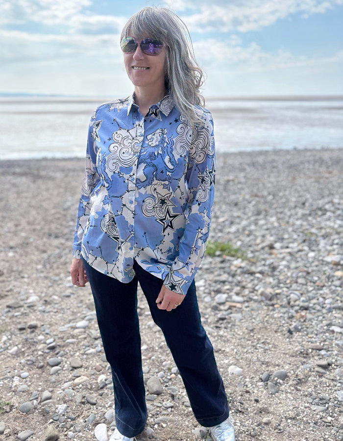 Person wearing a blue patterned shirt on a pebbly beach with water and sky in the background