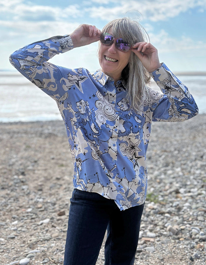 Person wearing a blue patterned shirt on a pebbly beach with ocean in the background