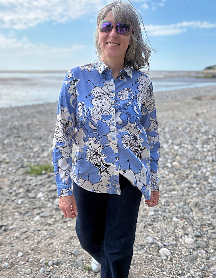 Person wearing a blue patterned shirt on a pebbly beach with a blue sky.