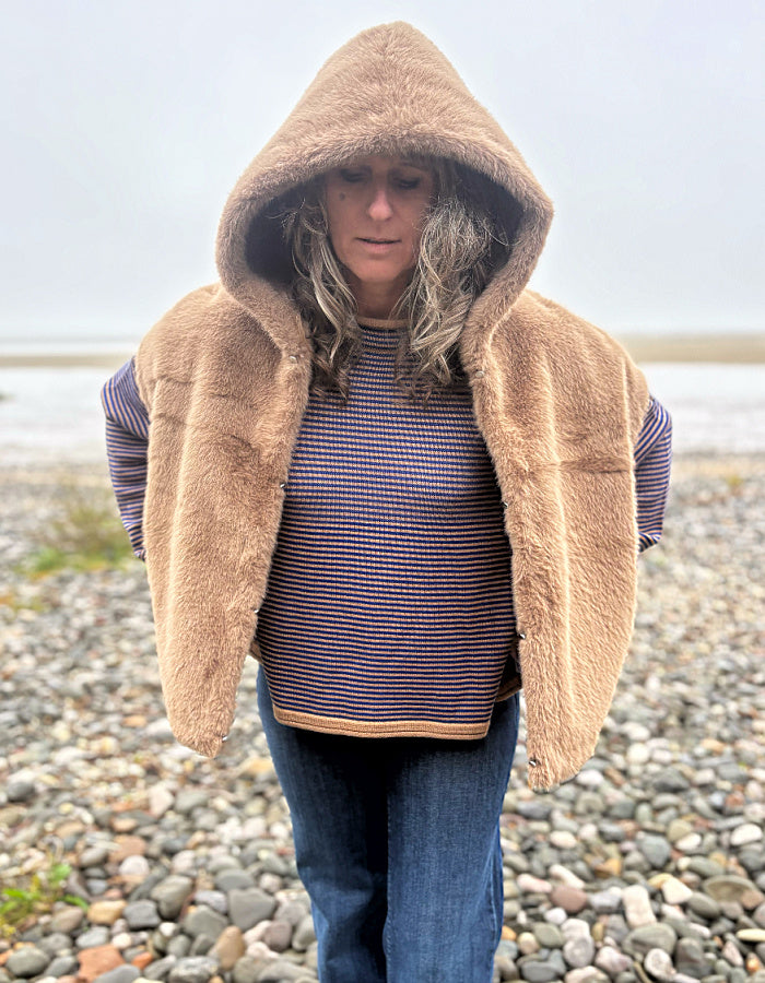 A woman standing on a pebble beach wearing a tan fuzzy hooded vest over a blue and red striped sweater and blue jeans.