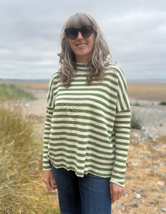 A woman with gray hair and sunglasses wearing a green and cream striped long-sleeve top, standing outdoors on a beach path.