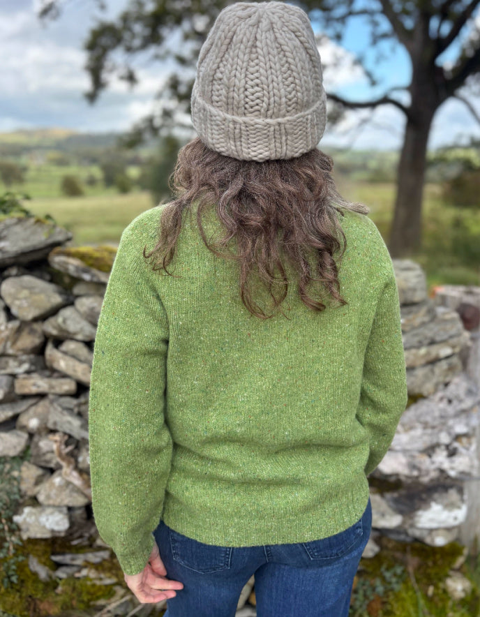 Person wearing a green knitted sweater and beige beanie standing in a natural setting with trees and stone wall.