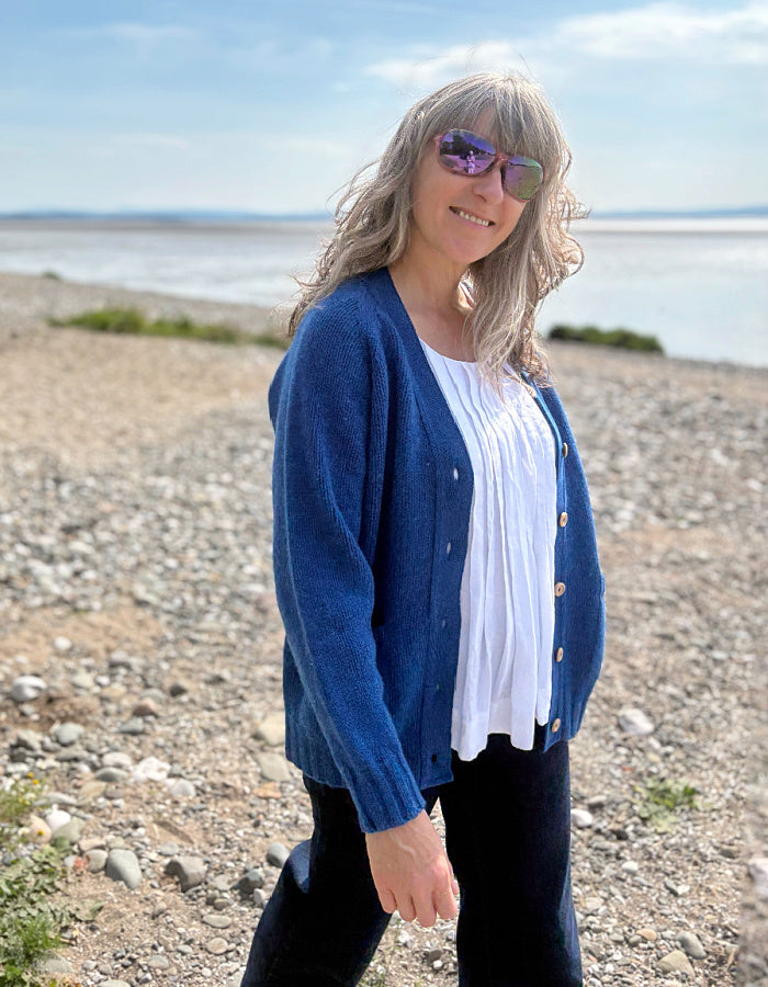 Woman wearing a wool blue cardigan on a pebbly beach with water and sky in the background