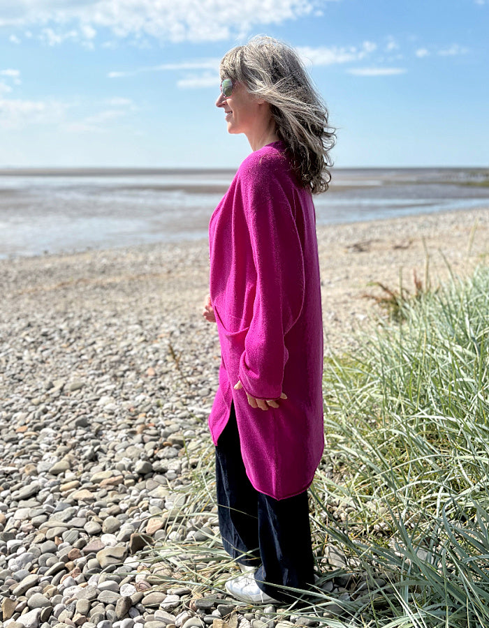 Person wearing a bright pink long wool cardigan standing on a pebbly beach with grass and water in the background.