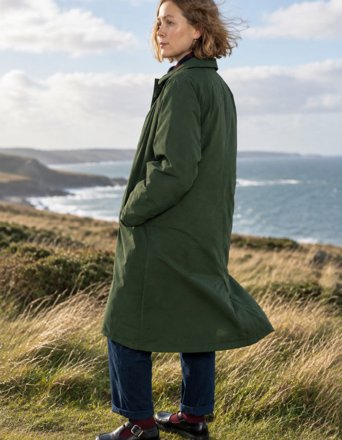 A woman wearing a long dark green coat, blue jeans, and brown shoes stands on a grassy coastal hillside with the ocean and cloudy sky in the background.