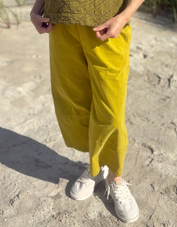 A person outdoors wearing loose mustard yellow cropped pants, a black-and-white checkered top, and white sneakers on sandy ground.