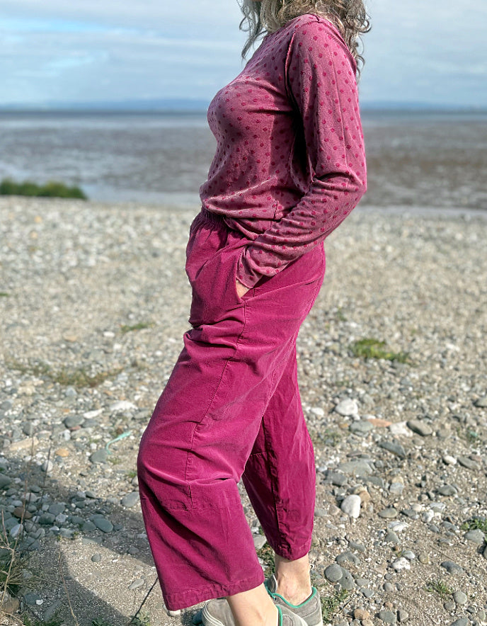 Woman in pink needlecord lantern trousers and a pink pink spotty top standing on a pebbly beach with ocean and sky in the background