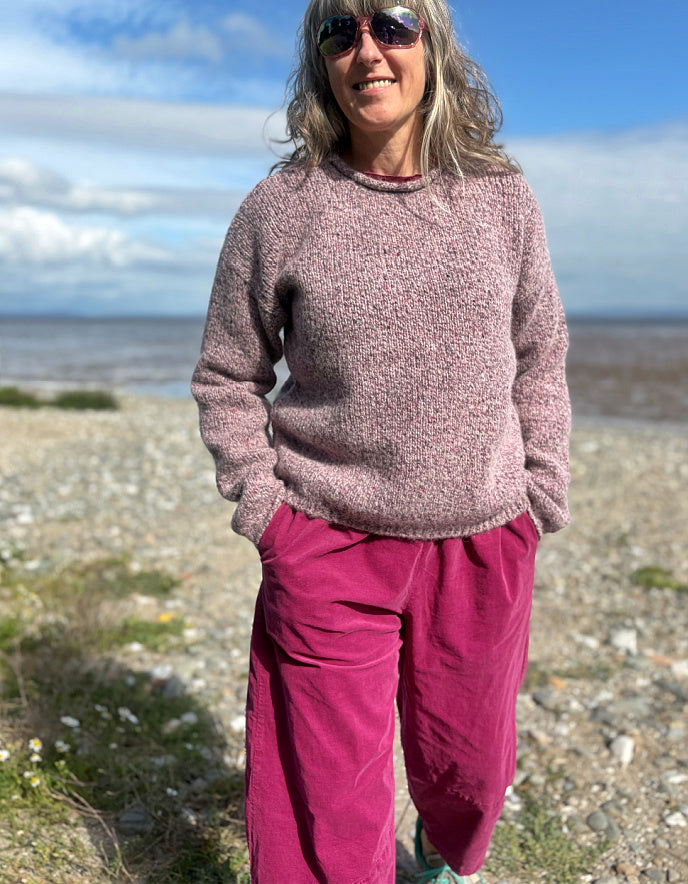 Woman standing on a beach wearing a pink sweater and purple pants, with sunglasses on.