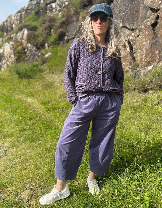 A woman stands outdoors on a grassy hillside with rocks in the background, wearing a quilted dark purple cropped cardigan, matching wide-leg purple pants, a blue cap, sunglasses, and white sneakers.